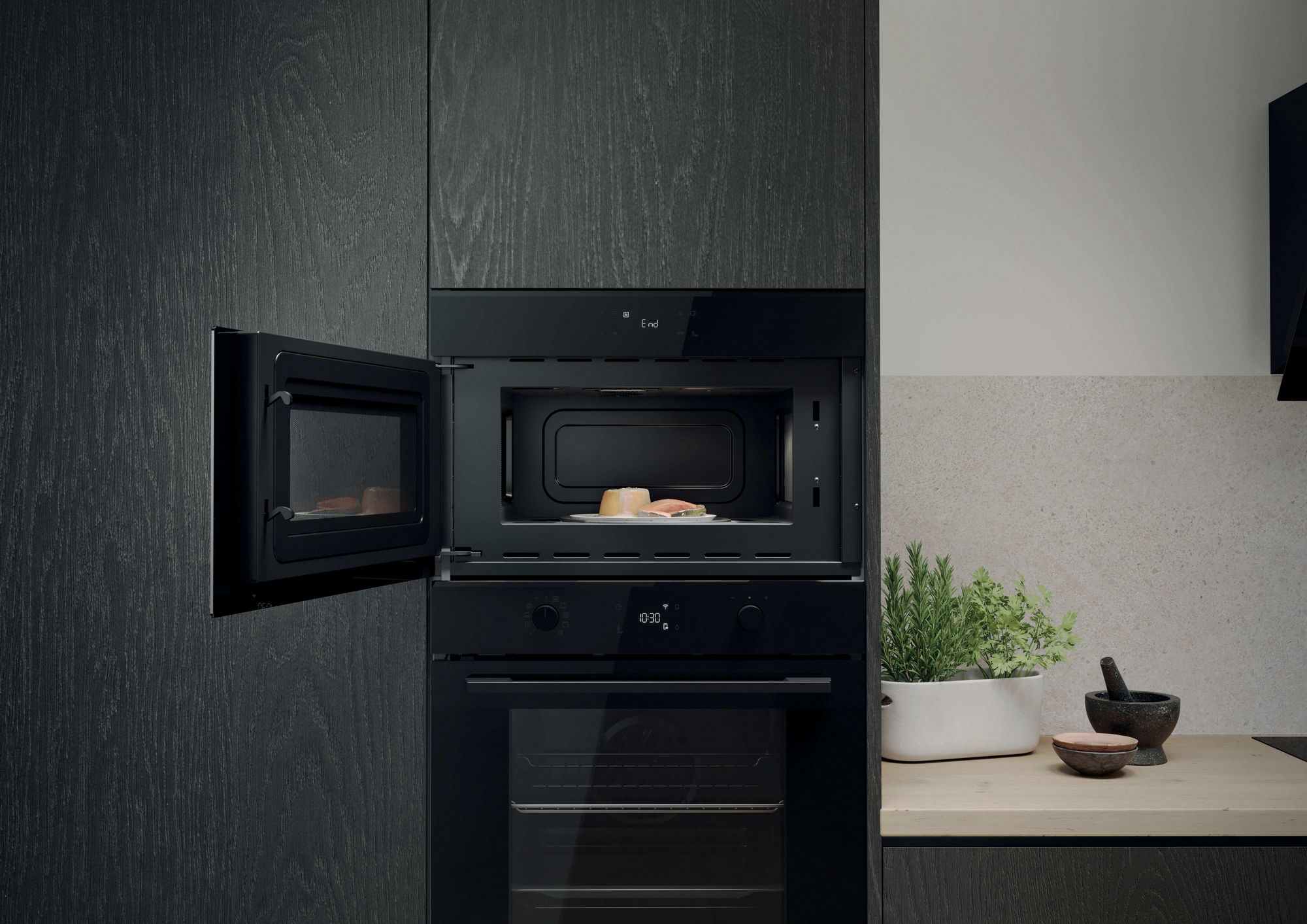 Combinación de microondas y horno negros con la puerta del microondas abierta. Comida dentro. Hierbas en una maceta blanca en la encimera.