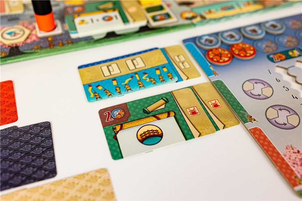 Widoczne elementy gry planszowej. Karty z ilustracjami, żetony i plansza. Kolory to beżowy, zielony i niebieski.