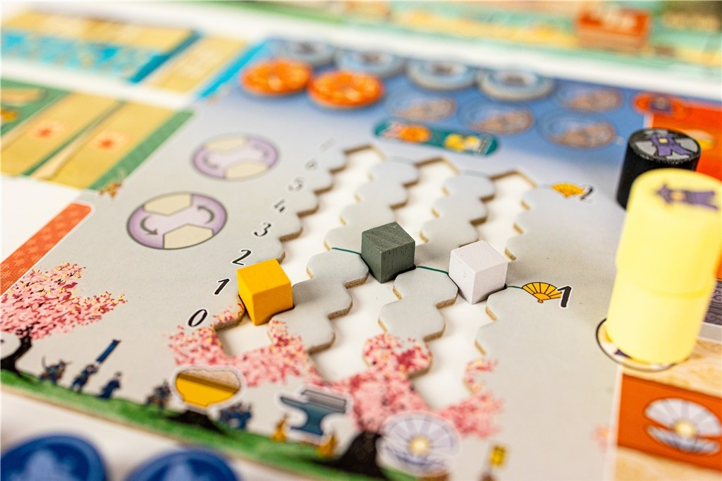 Gra planszowa z kolorowymi elementami. Trzy kostki: żółta, szara, biała. Widoczny jest również różowy wzór kwiatu wiśni i inne elementy.
