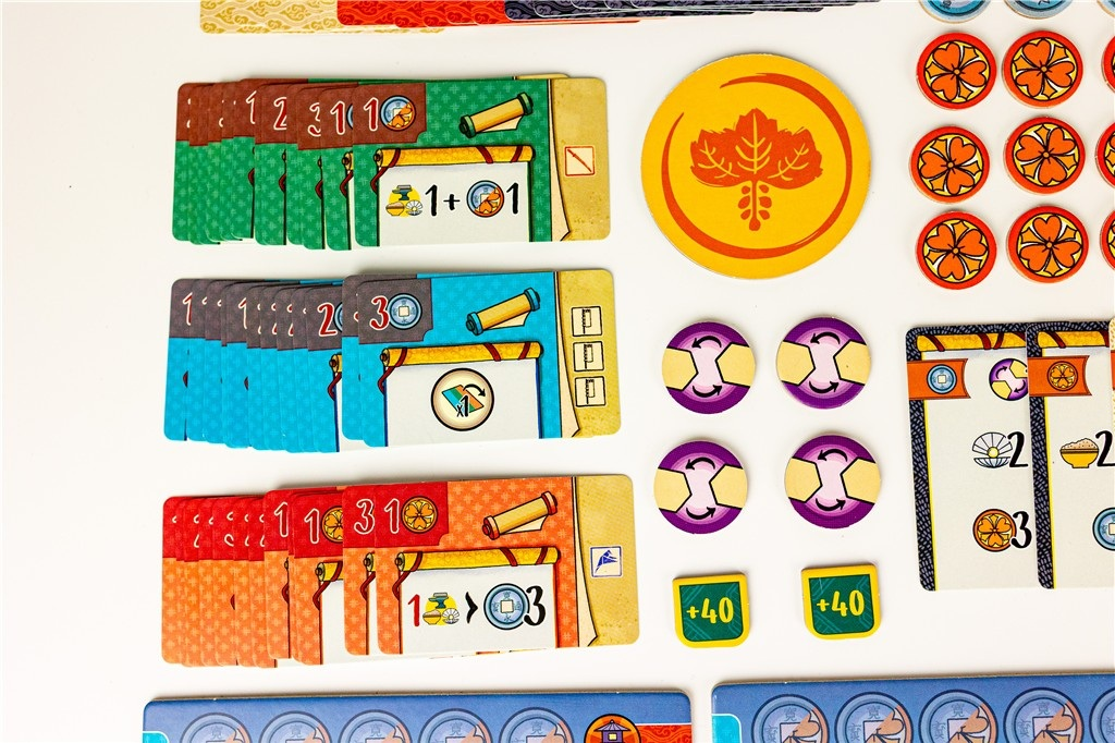 Elementy gry planszowej: karty, żetony i okrągłe elementy z różnymi symbolami i numerami, na białej powierzchni.