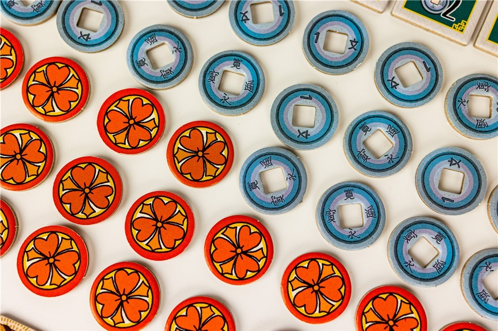 Widok z góry na elementy gry: czerwone żetony kwiatowe i niebieskie okrągłe żetony z japońskimi znakami i numerami, na białej powierzchni.