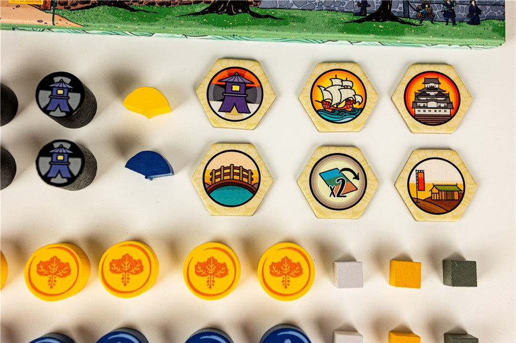 Elementy gry planszowej: żetony, sześciokątne płytki ze zdjęciami budynków i statków oraz małe drewniane kostki.
