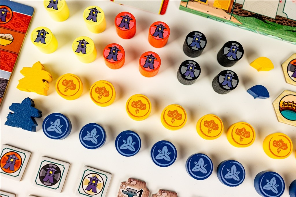 Elementy gry planszowej: kolorowe drewniane elementy, żetony i figurki graczy na białej powierzchni, widoczna plansza.