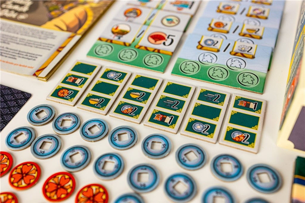 Elementy gry planszowej: karty, żetony i monety ułożone na białej powierzchni. Gra wydaje się mieć azjatycki motyw.