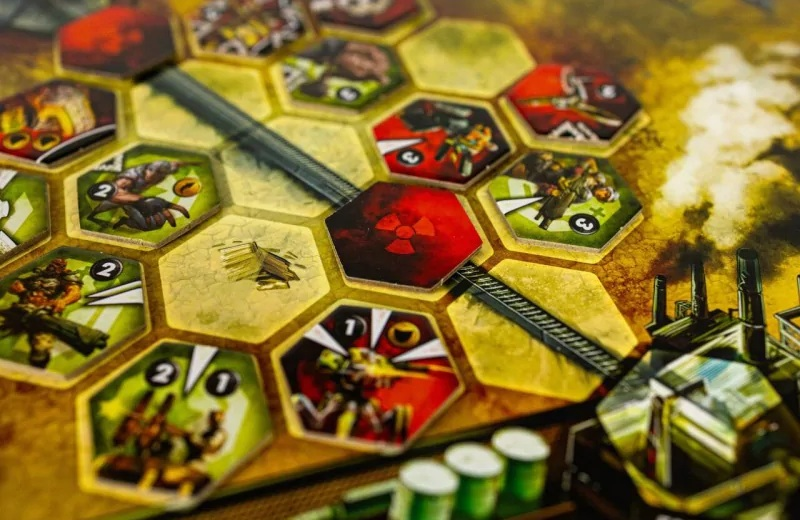 Gra planszowa z sześciokątnymi kafelkami. Czerwony kafelek z symbolem promieniowania. Postacie w scenach akcji, tło przemysłowe. Kolory zielony, żółty i czerwony.