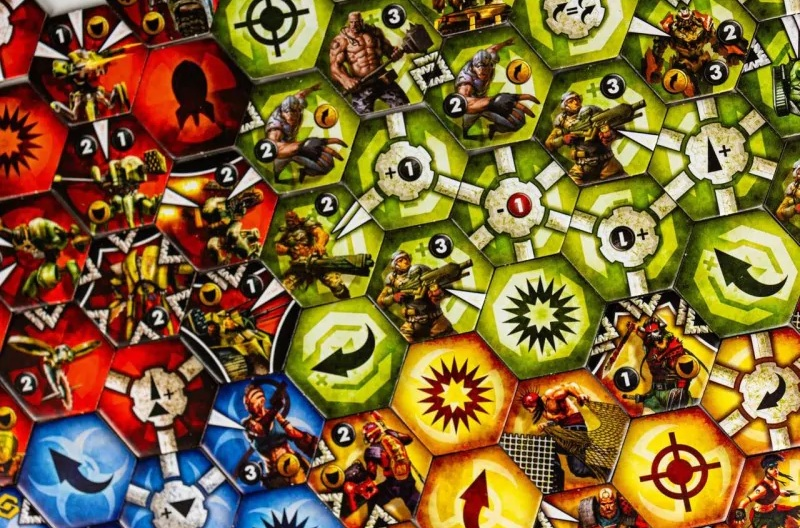 Gra planszowa z sześciokątnymi kafelkami. Kafelki zawierają ilustracje żołnierzy, broni i symboli w różnych kolorach, takich jak czerwony, zielony i żółty.