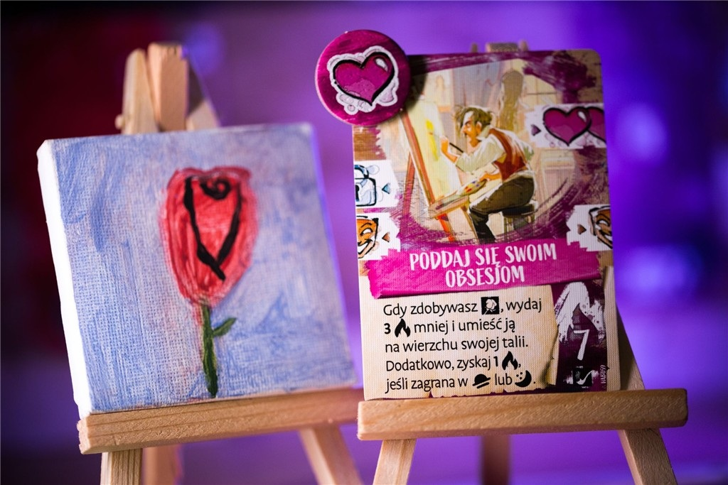 Dwa sztalugi prezentują sztukę: obraz róży i kartę z ilustracją artysty. Żeton w kształcie serca spoczywa na karcie.