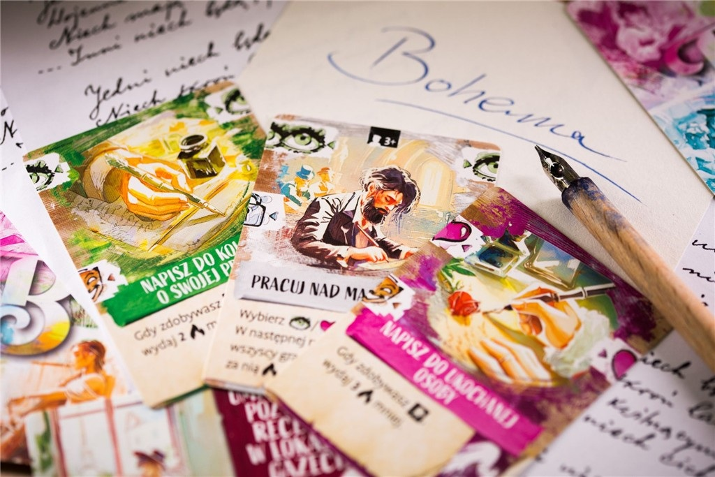Karty z ilustracjami i tekstem leżą na biurku z piórem i odręcznymi notatkami. Słowo 'Bohema' jest napisane na papierze.