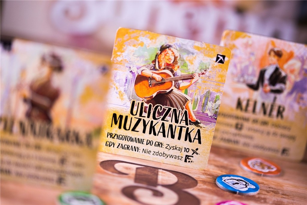 Karty z ilustracjami muzyka grającego na gitarze i kelnera. Żetony gry rozrzucone na drewnianej powierzchni.