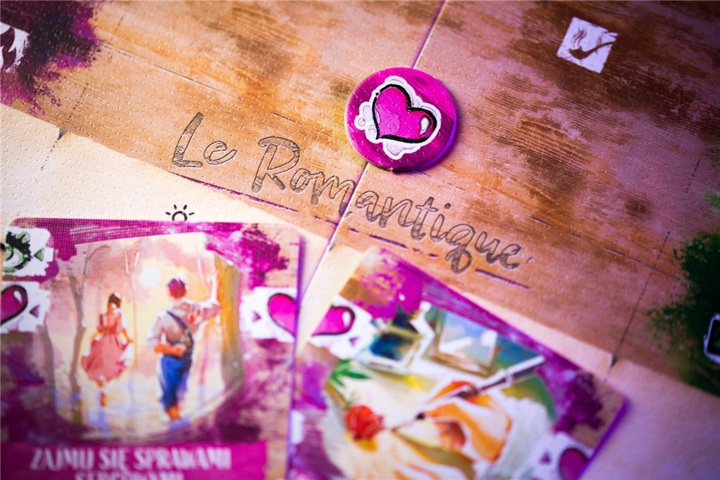 Gra planszowa z kartami i żetonem. Karty przedstawiają pary i artystów. Plansza mówi 'Le Romantique'. Różowy żeton z sercem.