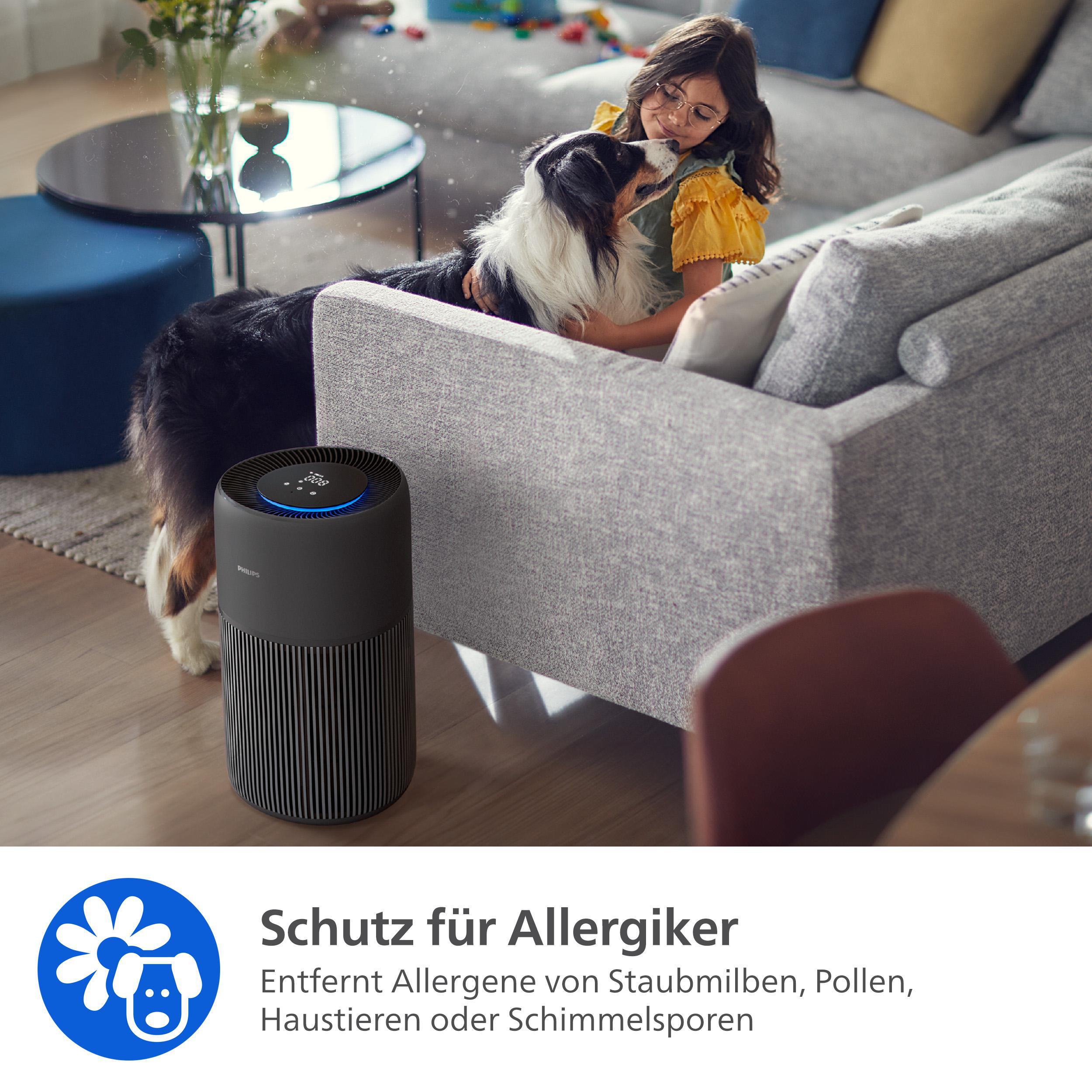 Ein Mädchen mit Brille streichelt einen Hund auf einem Sofa. Ein Luftreiniger befindet sich im Vordergrund. Blumen und ein Tisch sind im Hintergrund.