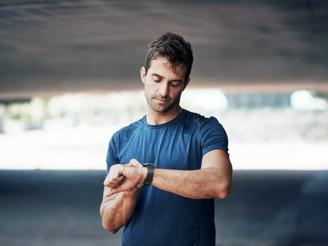Un homme en chemise bleue regarde sa montre sous un pont. Il regarde son poignet. L'arrière-plan est flou.