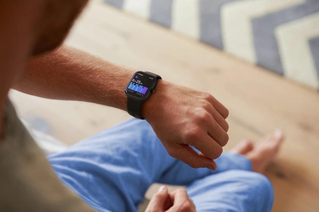 Une personne portant une smartwatch affichant des données de sommeil. La personne est assise, portant un pantalon bleu. Un plancher en bois et un tapis sont en arrière-plan.