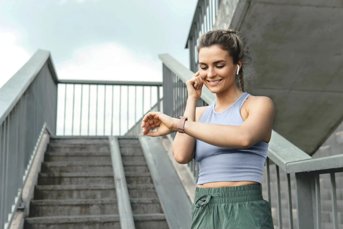 Femme en tenue de sport, souriante, regardant sa montre connectée. Escalier et structure en béton en arrière-plan.