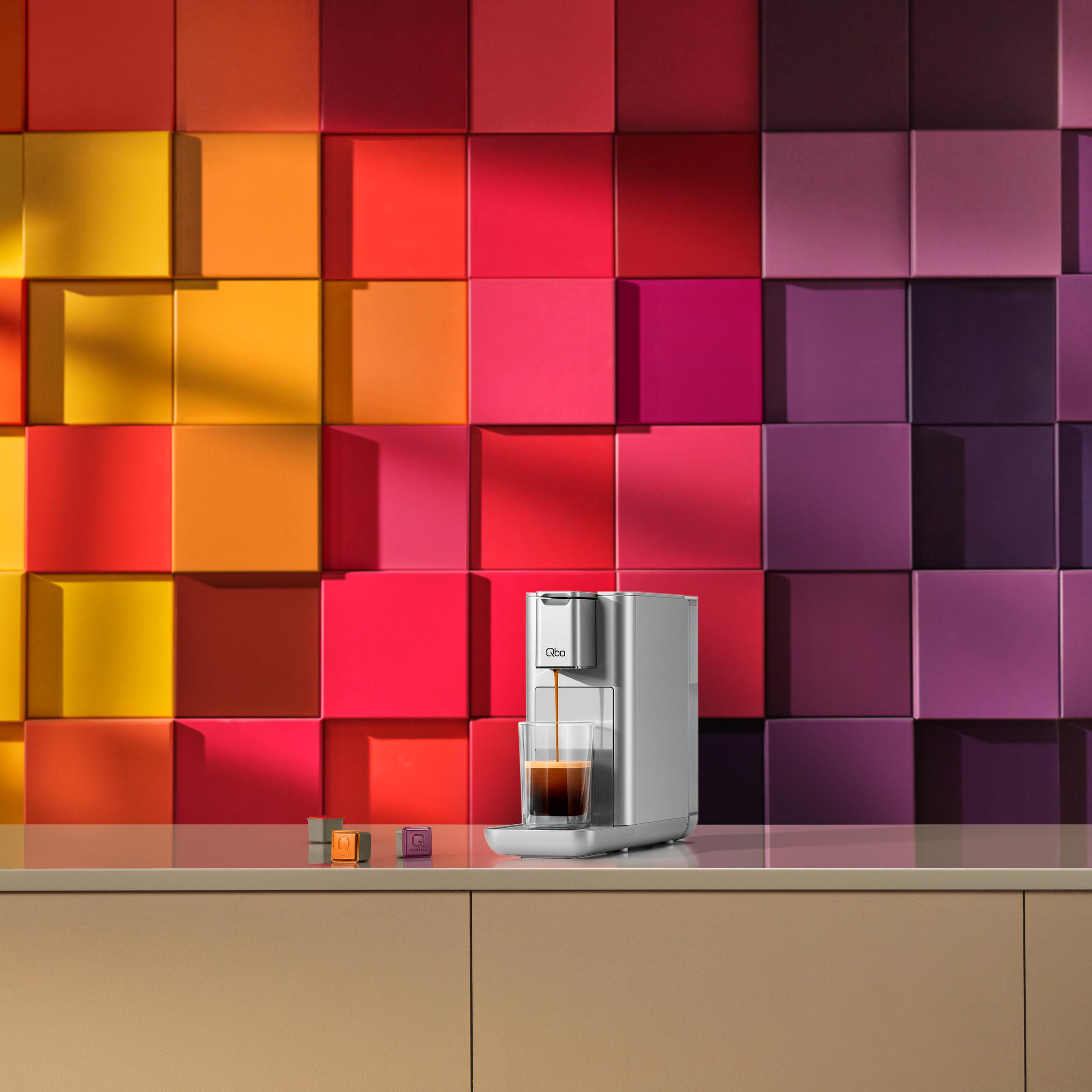 Silberne Qbo-Kaffeemaschine, die Espresso in ein Glas ausgibt. Bunter quadratischer Hintergrund, Kaffeepads auf der Theke.