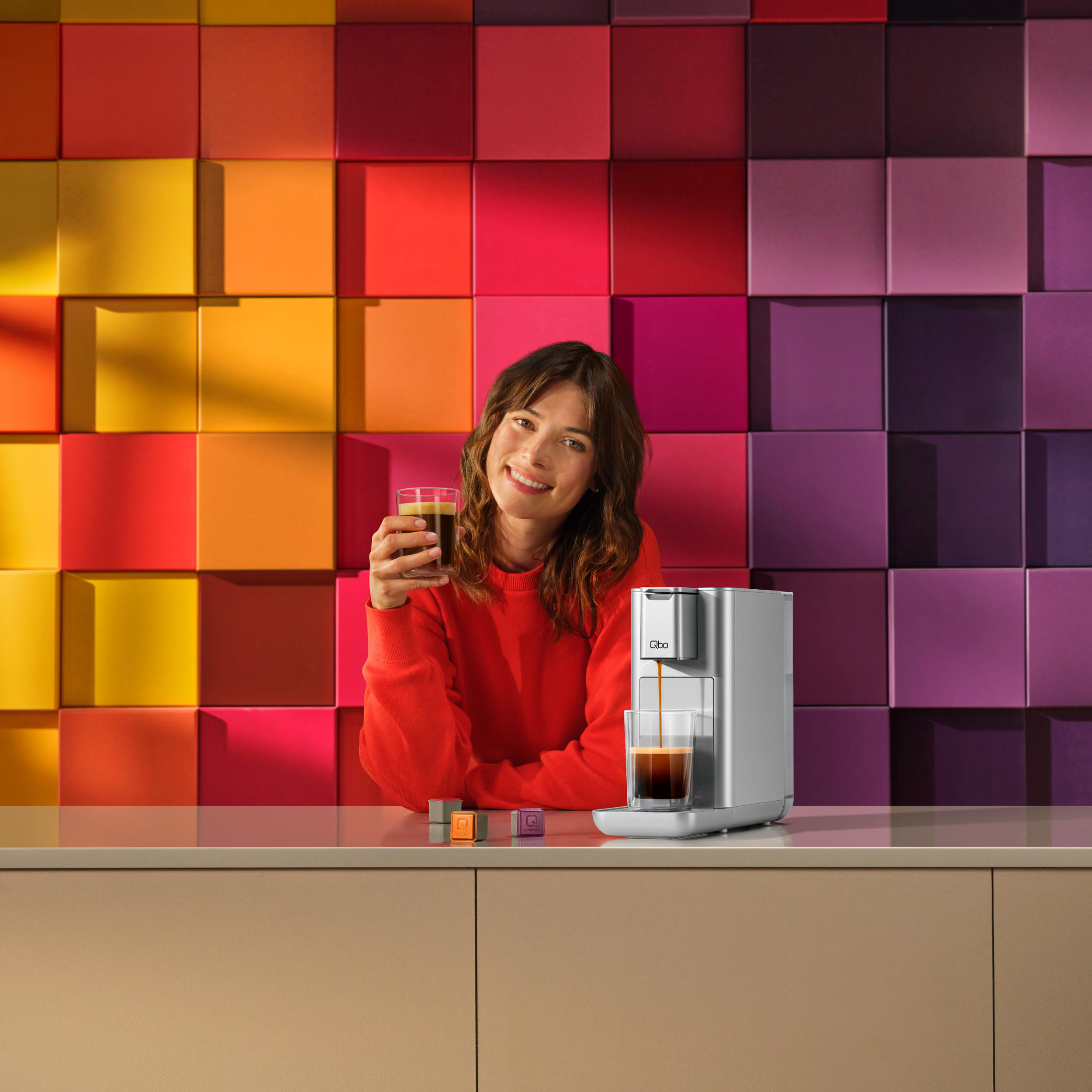 Frau hält einen Kaffee. Eine Kaffeemaschine steht rechts. Bunte quadratische Fliesen im Hintergrund.
