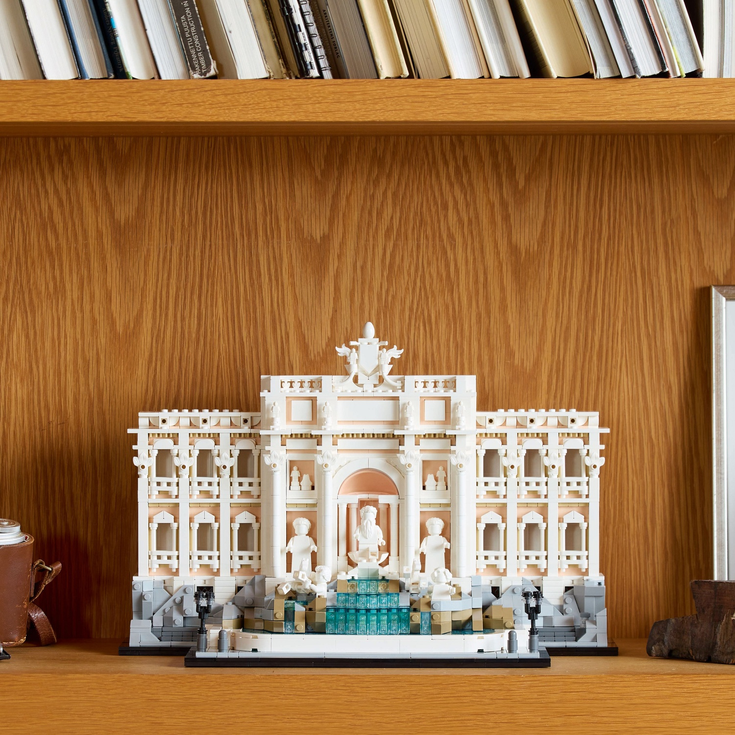 Een Lego-model van een gebouw staat op een houten plank. Bovenop staan boeken. Het model heeft witte, beige en grijze elementen.
