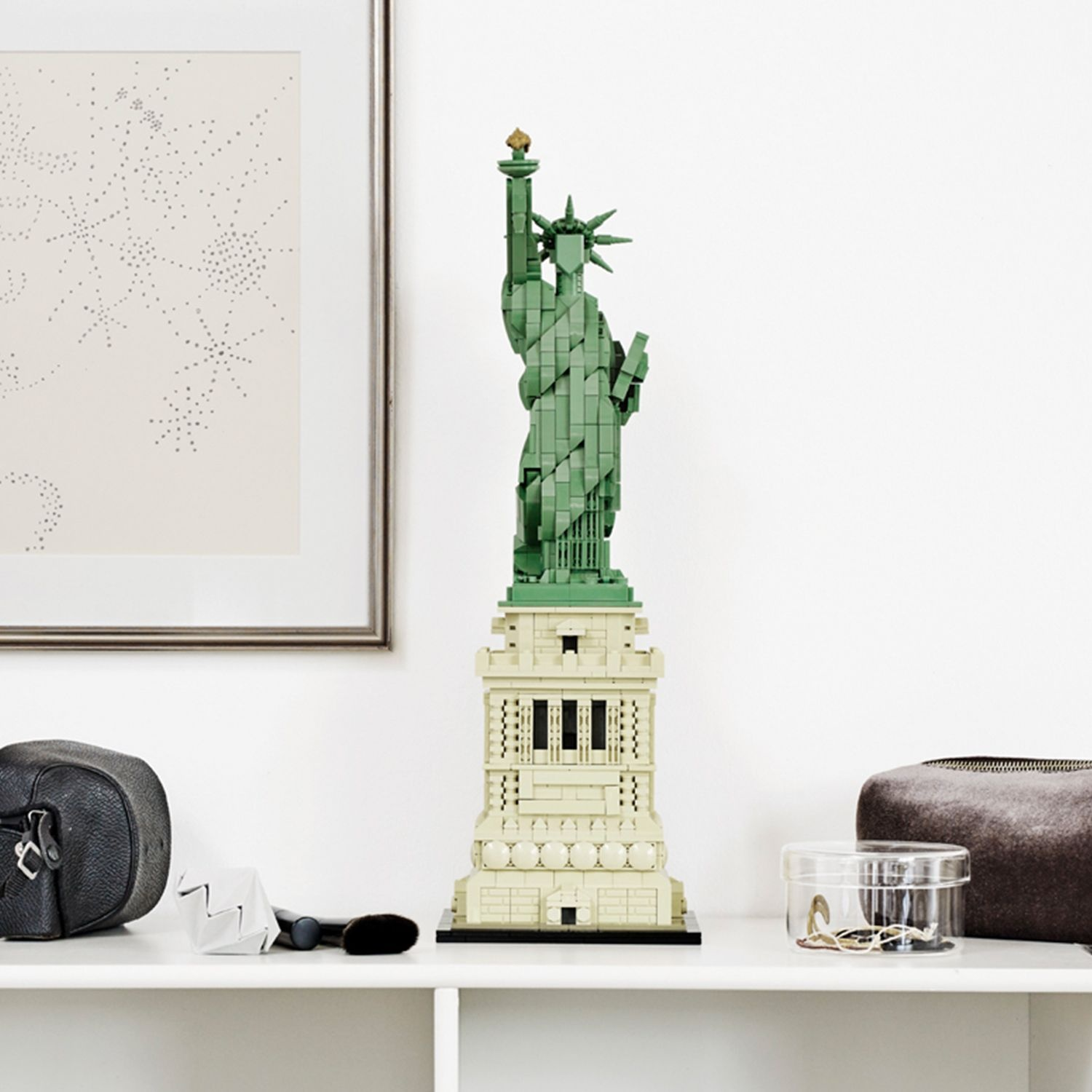 Une statue de la Liberté Lego est sur une étagère. Des objets comme un sac, une brosse et une boîte sont également présents. Une œuvre d'art encadrée est suspendue au-dessus.