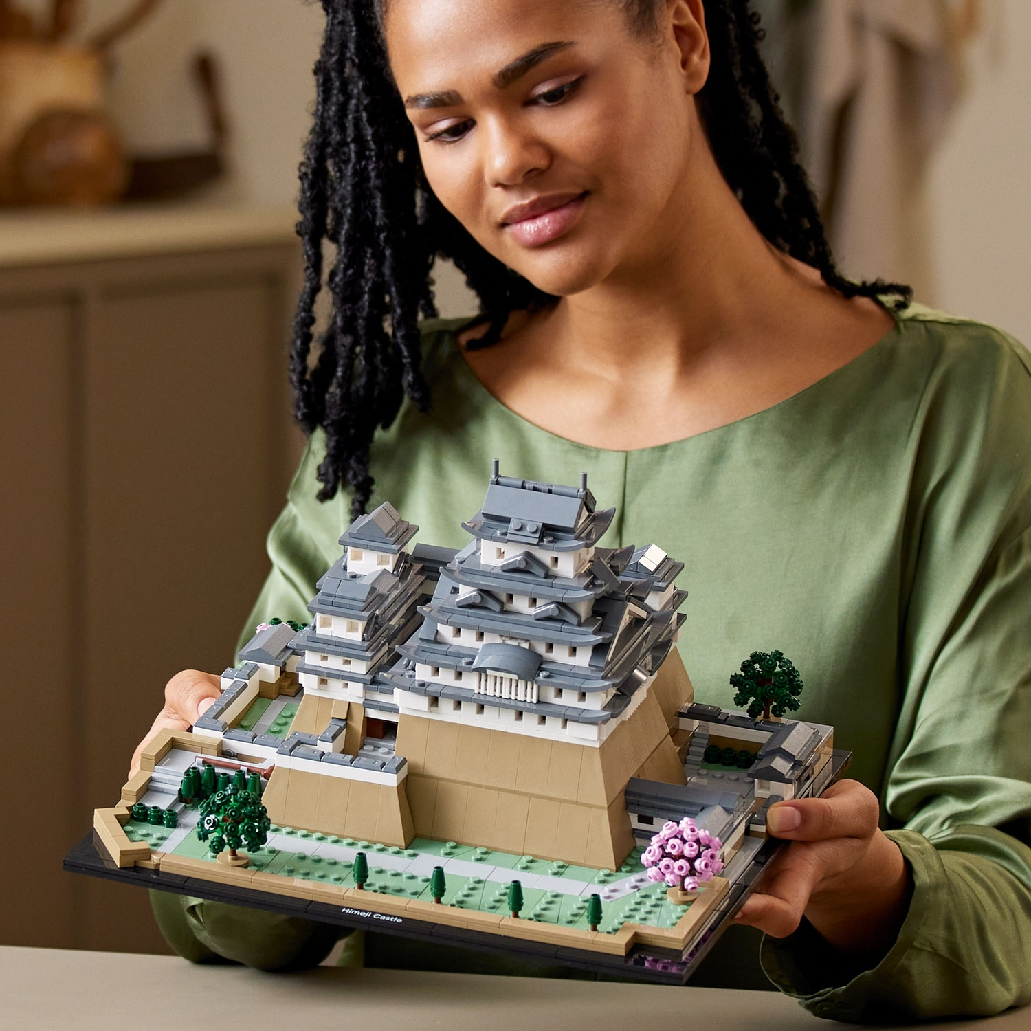 Een vrouw houdt een Lego-model van Kasteel Himeji vast. Het model is gedetailleerd met grijze daken, beige muren en groene omgeving.