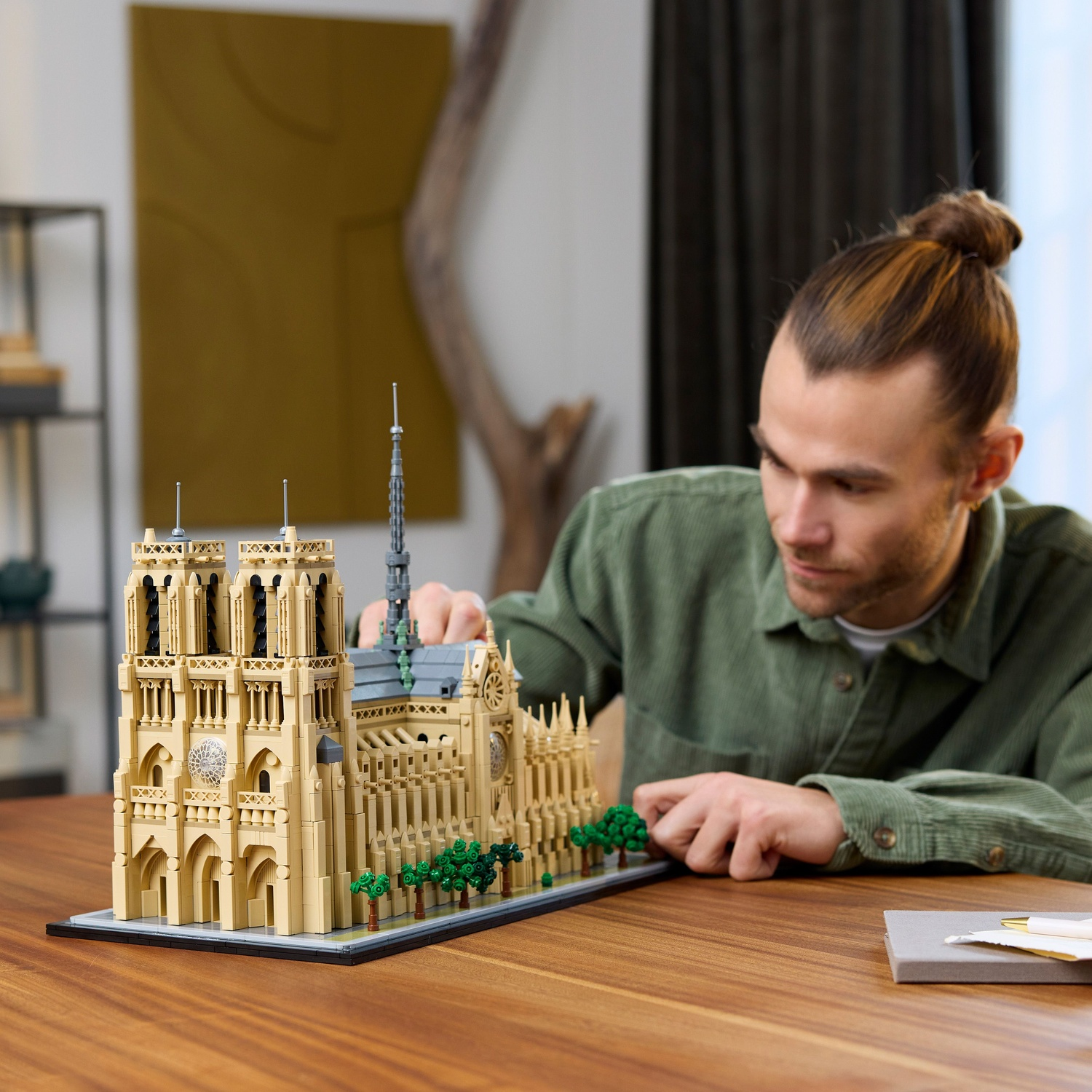 Een man bekijkt een Lego Notre Dame model op een houten tafel. Het model is beige, met groene bomen. Er ligt ook een notitieboek en een pen op de tafel.