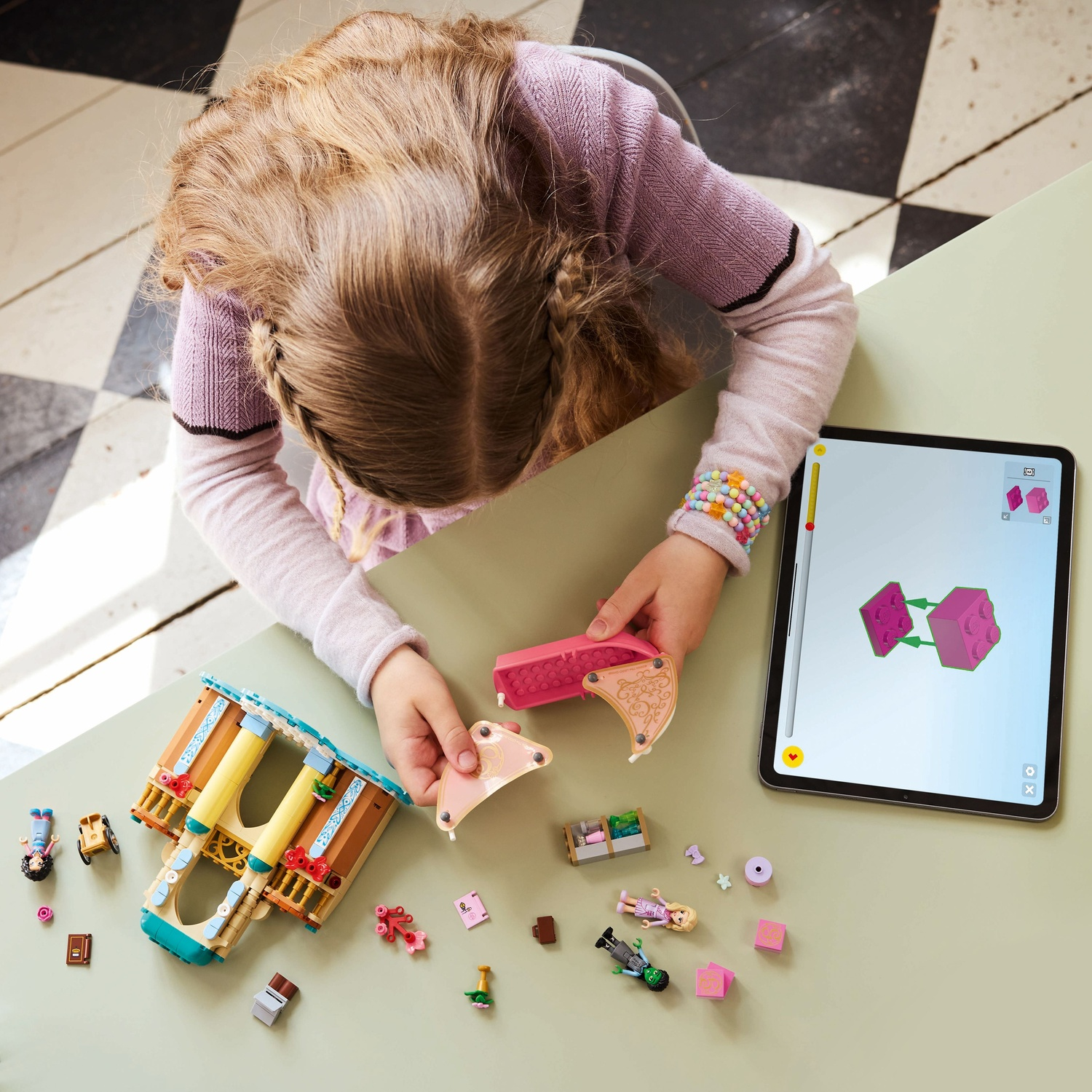 Een kind bouwt een speelgoedconstructie op een tafel. Lego-stukken liggen verspreid. Een tablet toont bouwinstructies.