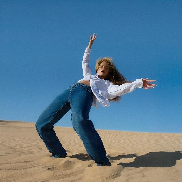 Eine Frau in Jeans und weißem Hemd tanzt im Sand unter blauem Himmel. Ihre Arme sind ausgestreckt, die Haare lockig.