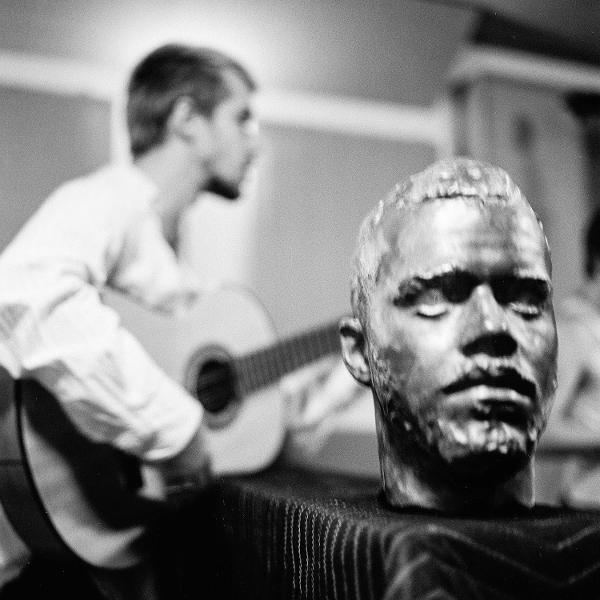 Eine Bronzebüste eines Mannes mit geschlossenen Augen. Im Hintergrund spielt ein Mann Gitarre. Schwarz-Weiß-Foto.