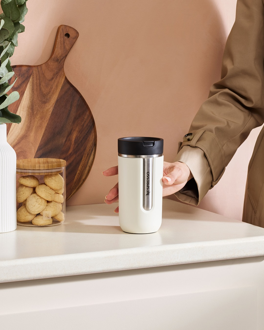 Beyaz bir yüzeyde bej renkli bir Nespresso seyahat kupası duruyor. Bir el ona uzanıyor. Kavanozda kurabiyeler ve sol tarafta bir tahta.