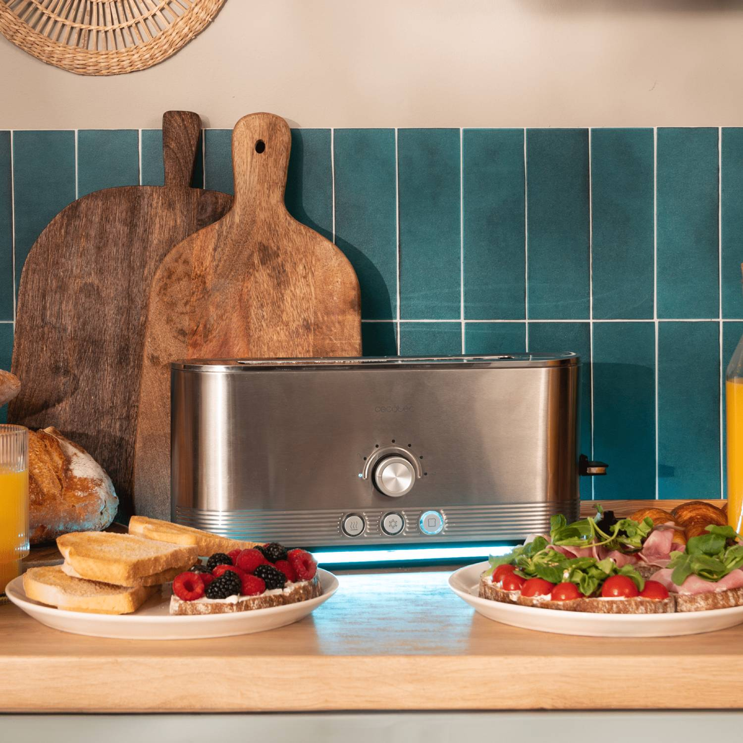 Una tostadora de acero inoxidable, dos platos de tostadas, fruta y sándwich, tablas de madera y zumo de naranja. Fondo de azulejos turquesa.