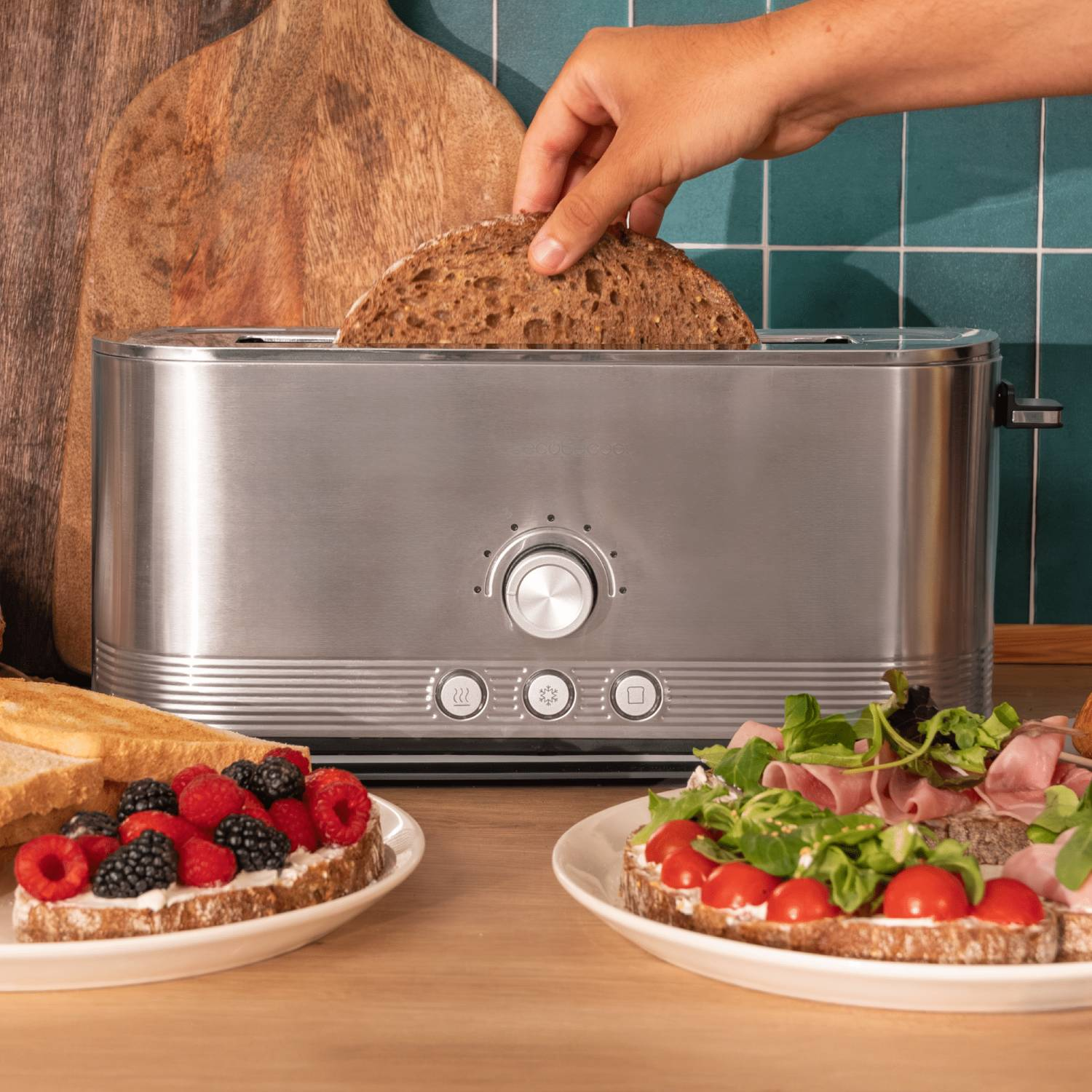 Una mano colocando pan en una tostadora. Dos sándwiches con bayas y jamón están en una mesa de madera. Una tabla de cortar está al fondo.