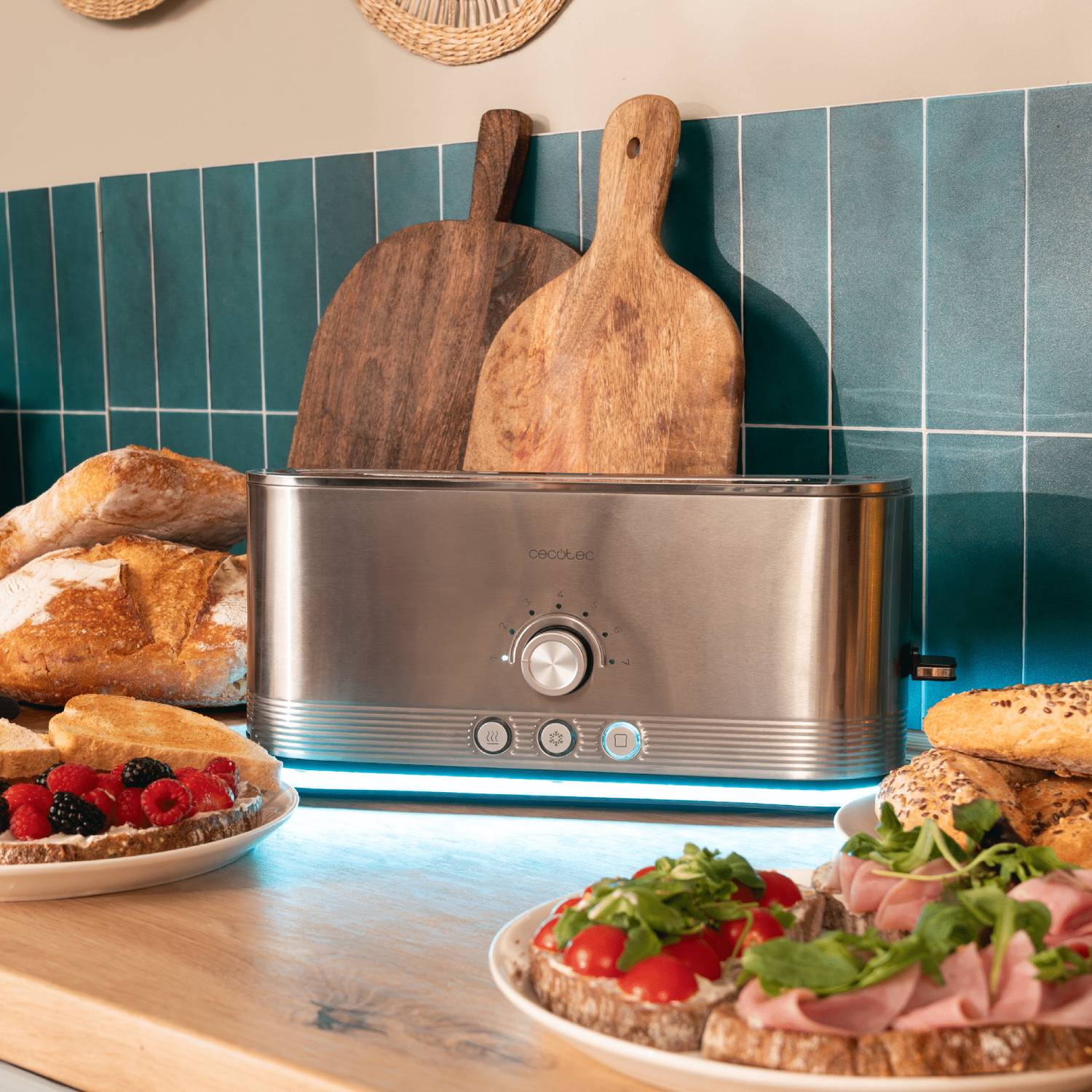 Tostadora de acero inoxidable con pan, sándwiches y tablas de cortar sobre una encimera de madera. Azulejos azules al fondo.