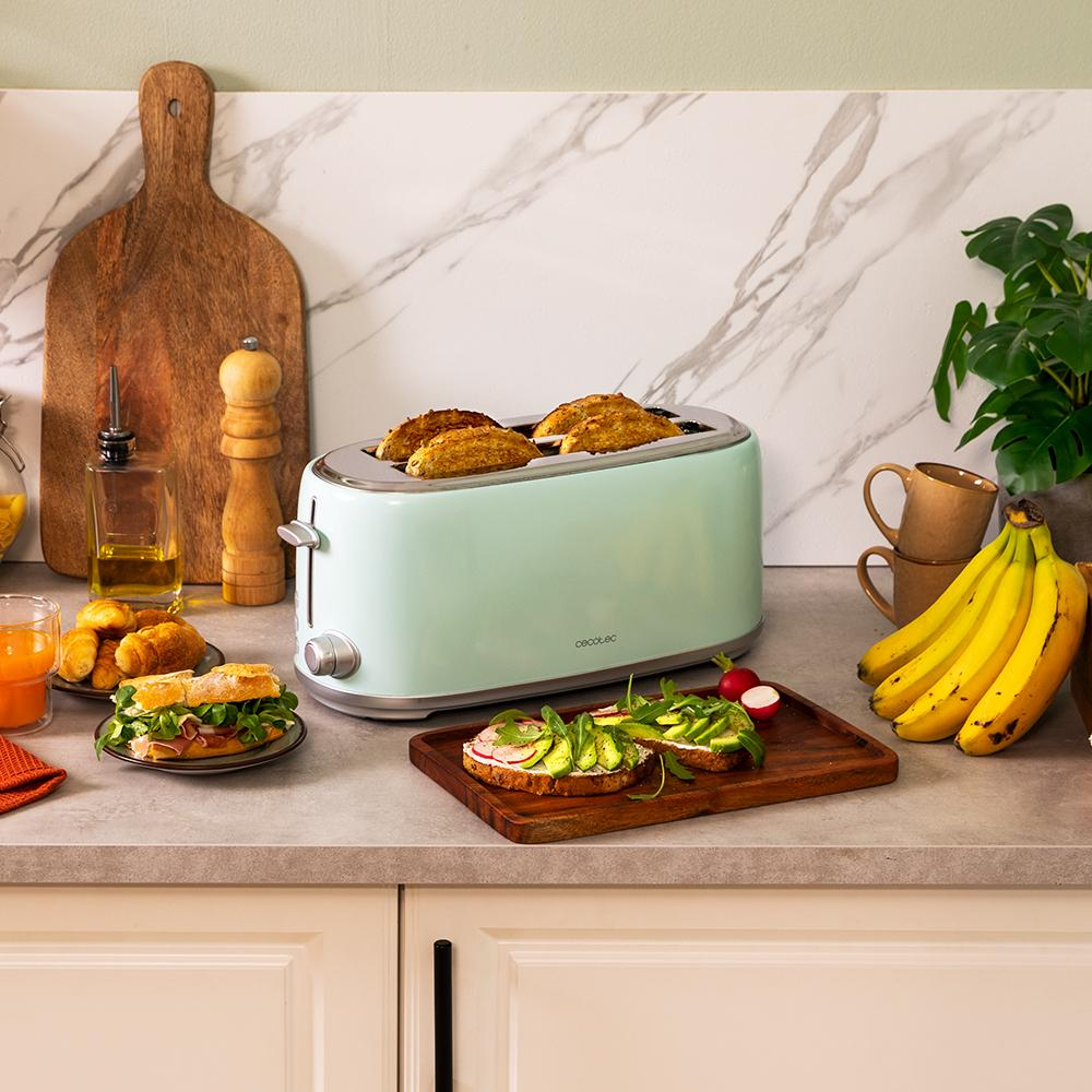 Una tostadora verde menta con pan tostado, sándwiches y una tabla de madera con tostadas de aguacate, plátanos y tazas de café.