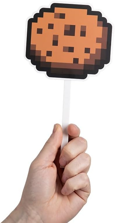 Egy kéz egy süti kivágást tart egy pálcán. A süti barna, sötétbarna pixeles részletekkel. A pálca fehér.