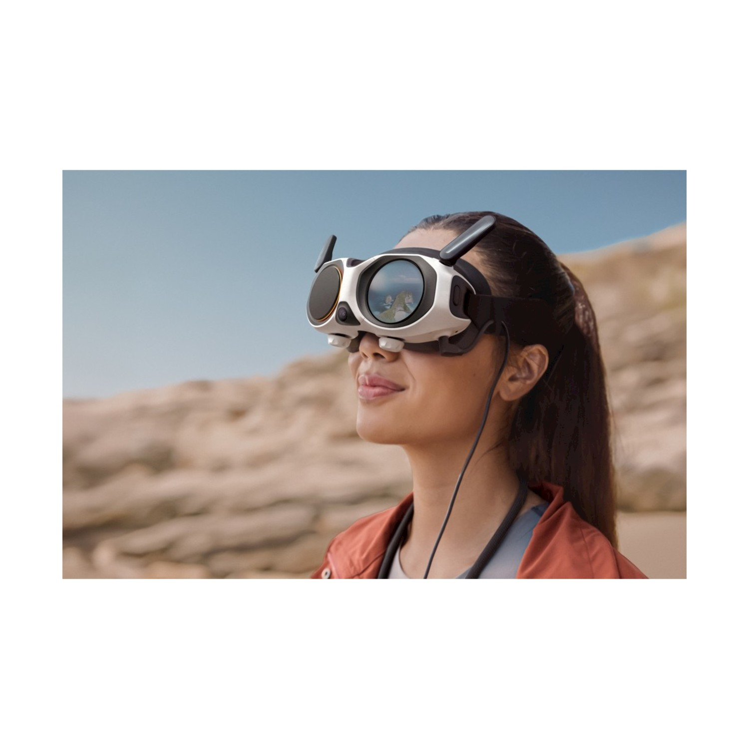 Una donna con occhiali VR, che guarda in alto. Gli occhiali riflettono un paesaggio. Indossa una giacca rossa, lo sfondo è un deserto sfocato.