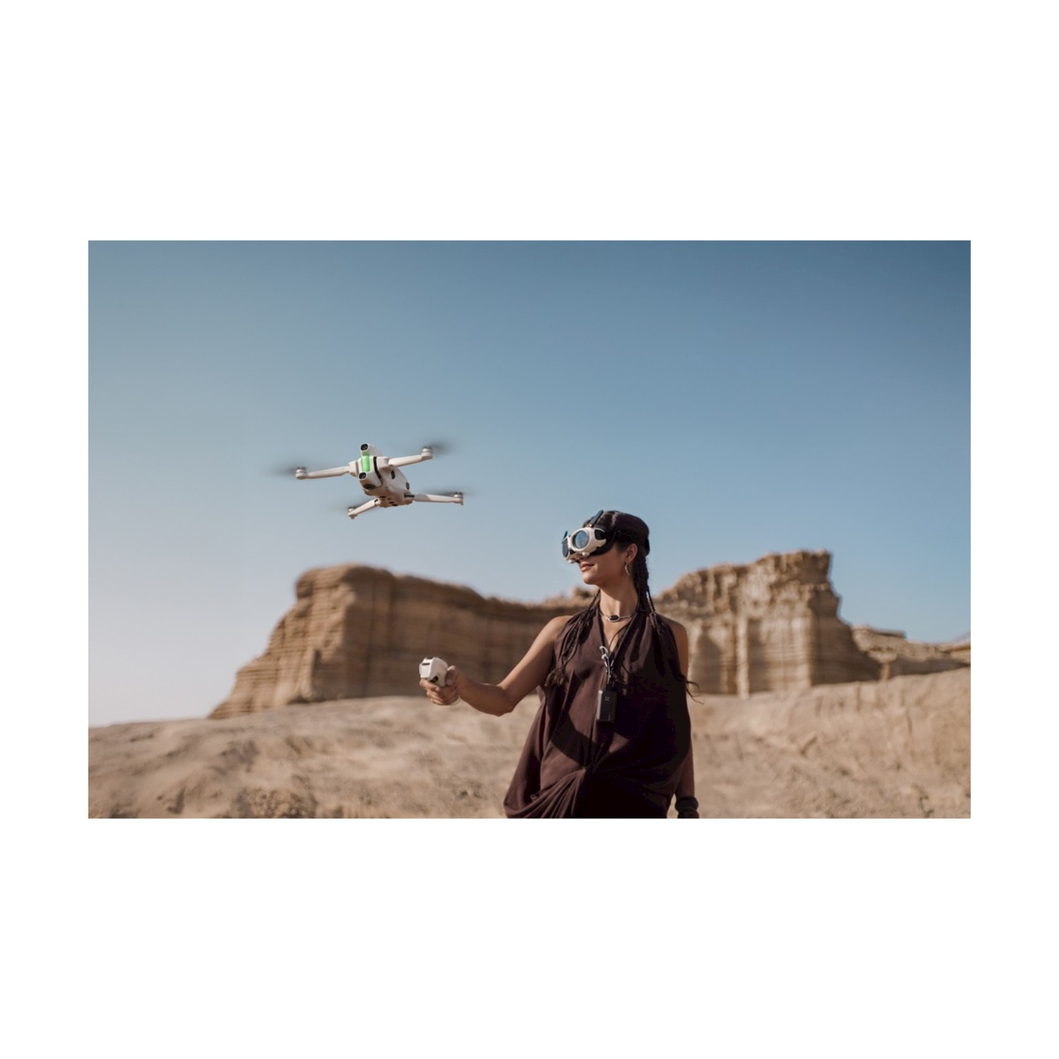 Una donna in un ambiente desertico fa volare un drone. Indossa occhiali e un abito scuro, tenendo in mano un controller. Il drone è in volo.