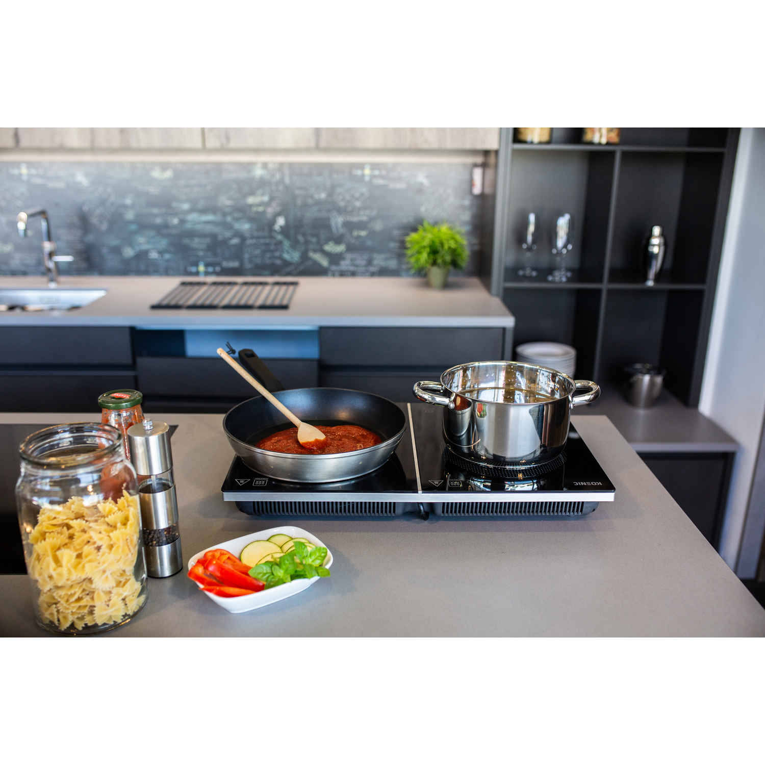 A kitchen scene with a pan of sauce and a pot on a cooktop. Pasta, spices, and vegetables are on the counter.