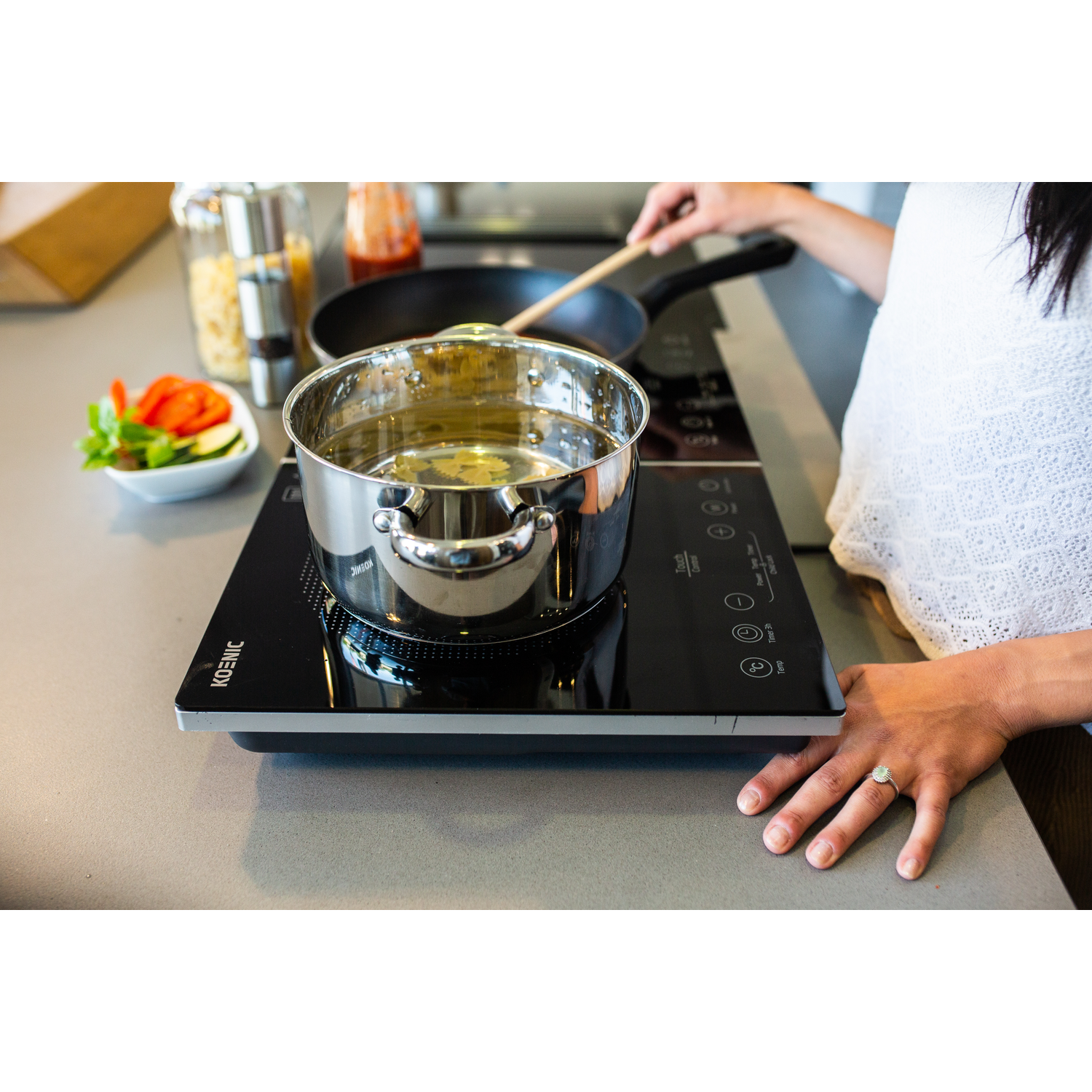 A woman cooks on a black induction cooktop. A pot of water boils. Ingredients sit nearby. A hand rests on the counter.