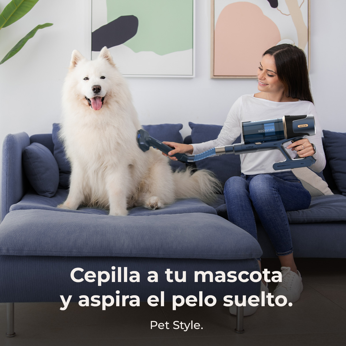 Una mujer aspira a un perro blanco y esponjoso en un sofá azul. La aspiradora es azul y negra. Texto: Cepille a su mascota y aspire el pelo suelto.