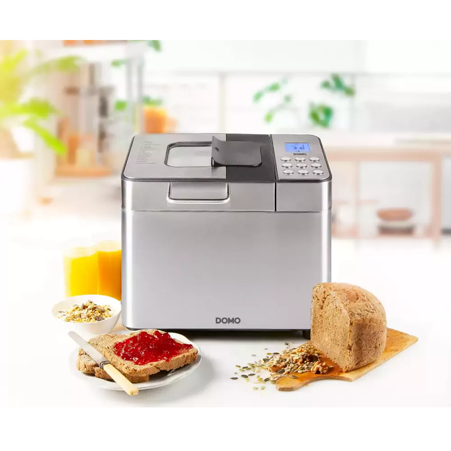 Een broodbakmachine met brood, toast met jam, sap en een kom ontbijtgranen staan op een tafel. De achtergrond is een vage keuken.