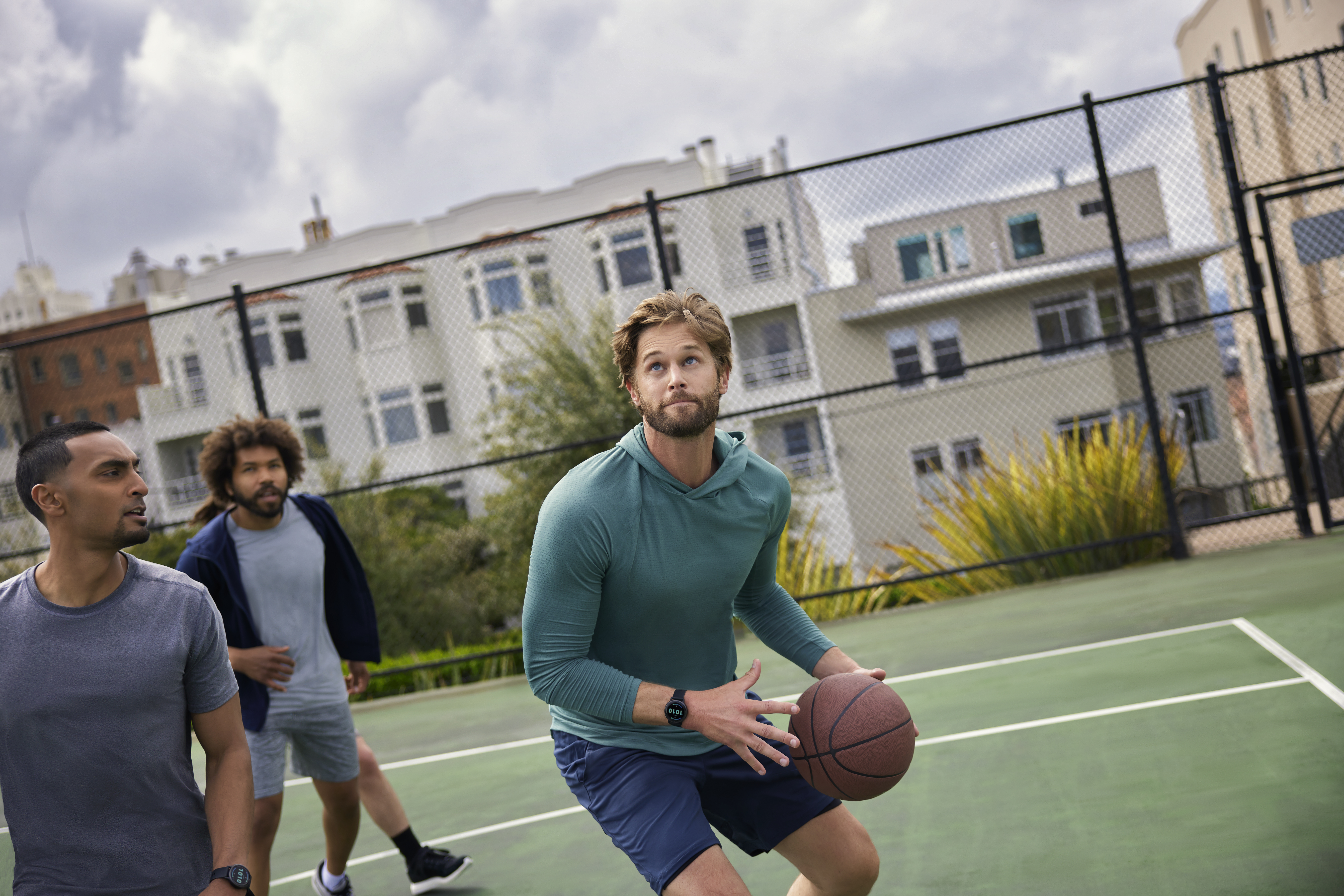Trzech mężczyzn gra w koszykówkę. Jeden trzyma piłkę, ubrany na zielono. Dwóch innych patrzy. Boisko z ogrodzeniem i budynkami w tle.