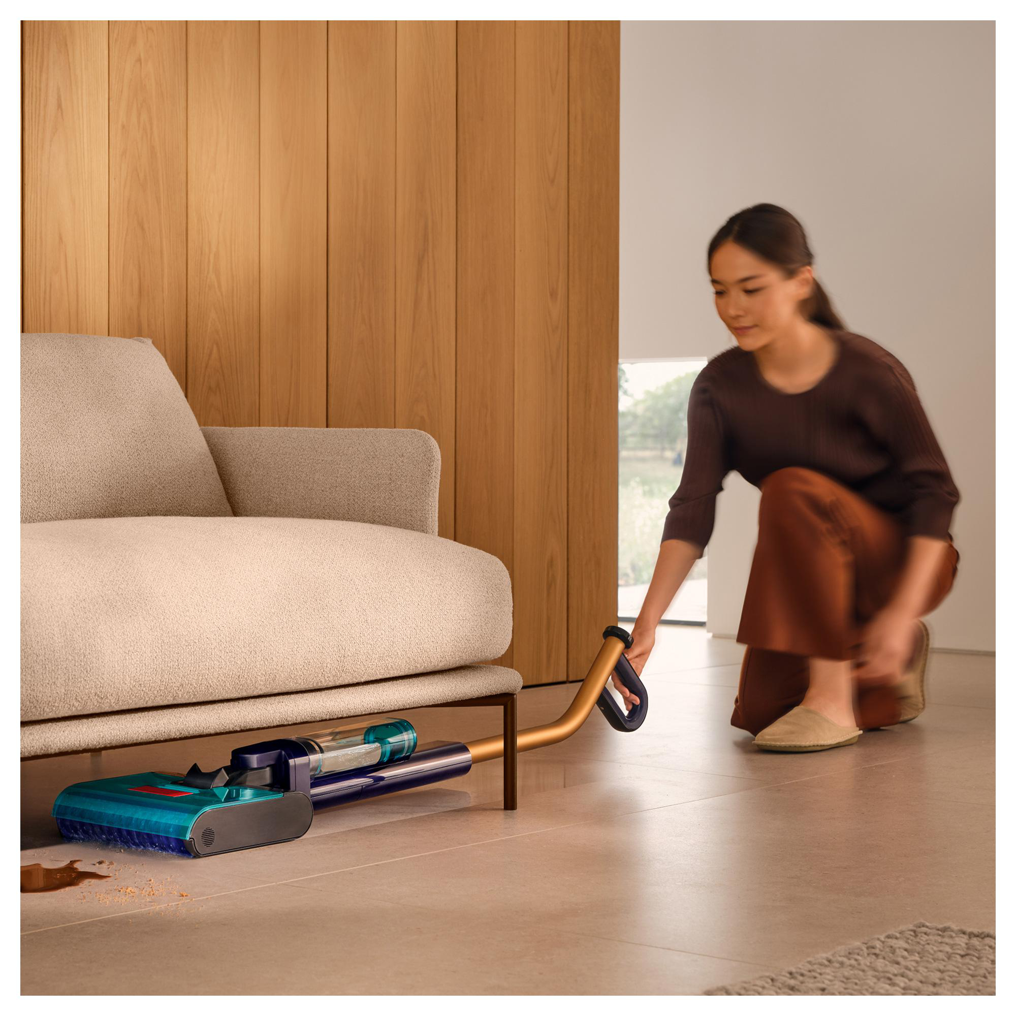 Frau saugt unter einem Sofa. Der Staubsauger ist türkis und lila. Brauner Fleck auf dem Boden. Holzwand im Hintergrund.