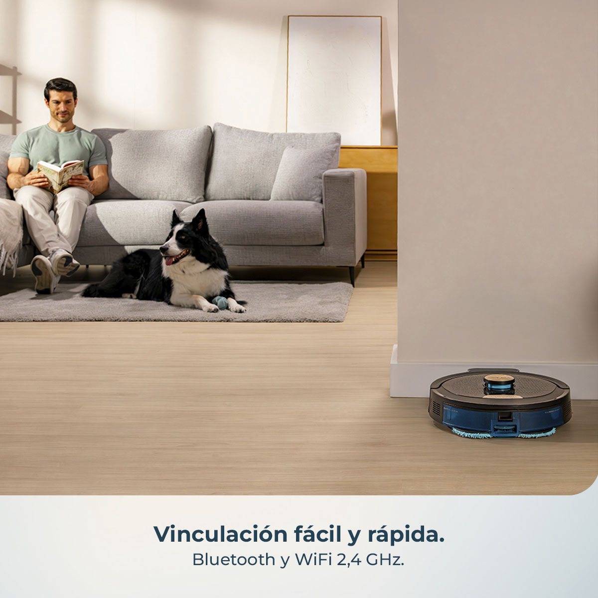 Un hombre lee un libro en un sofá con un perro. Una aspiradora robótica está en el suelo. La escena es en una sala de estar.