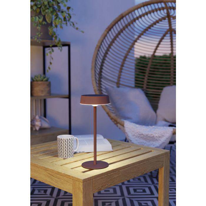 Eine Lampe auf einem Holztisch, eine Tasse und ein Buch. Im Hintergrund ein Korbsessel und Pflanzen.