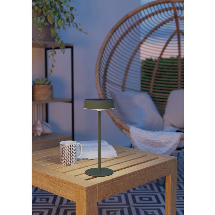 Eine Lampe auf einem Holztisch, mit einer Tasse und einem Buch. Ein Korbstuhl und Pflanzen sind im Hintergrund.