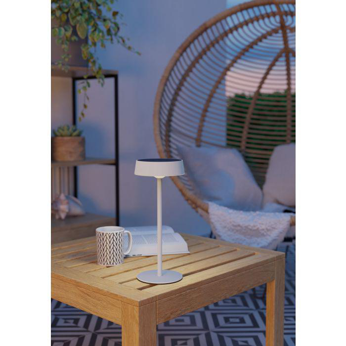 Eine weiße Lampe, eine Tasse und ein Buch stehen auf einem Holztisch. Ein Korbstuhl und Pflanzen sind im Hintergrund.