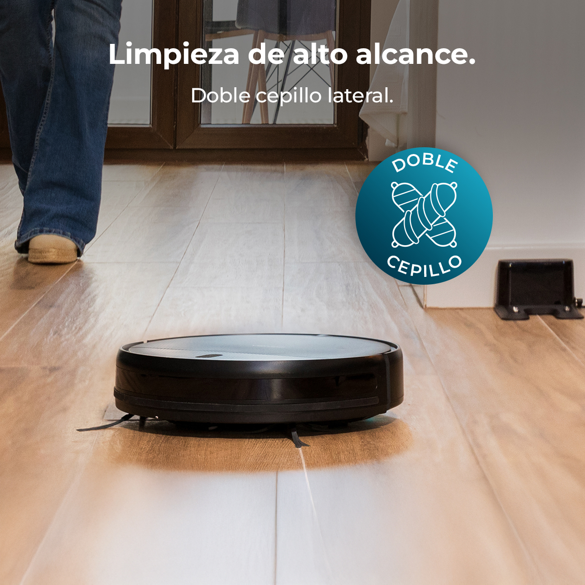 Una aspiradora robot negra en un piso de madera. Una persona con jeans y zapatos color canela camina atrás. Un cargador a la derecha.