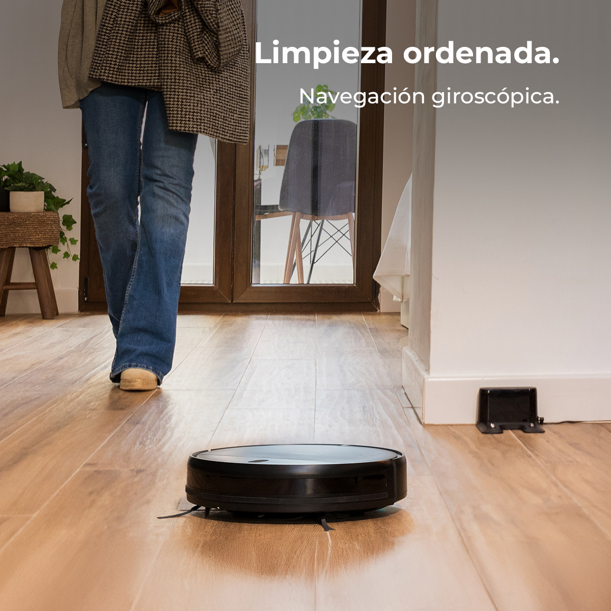 Una persona camina sobre piso de madera hacia una aspiradora robot negra. Una estación de carga está a la derecha. Espejo refleja una silla.