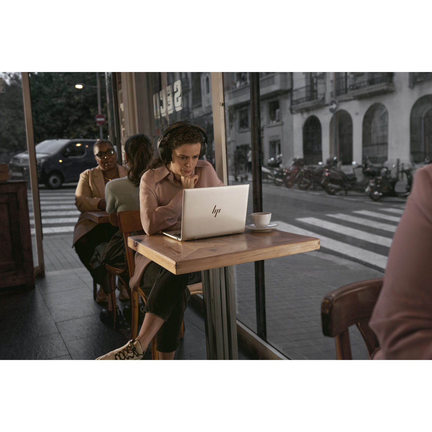 Vrouw met koptelefoon gebruikt laptop aan een cafétafel. Anderen zitten in de buurt. Auto's en motoren zijn buiten zichtbaar.