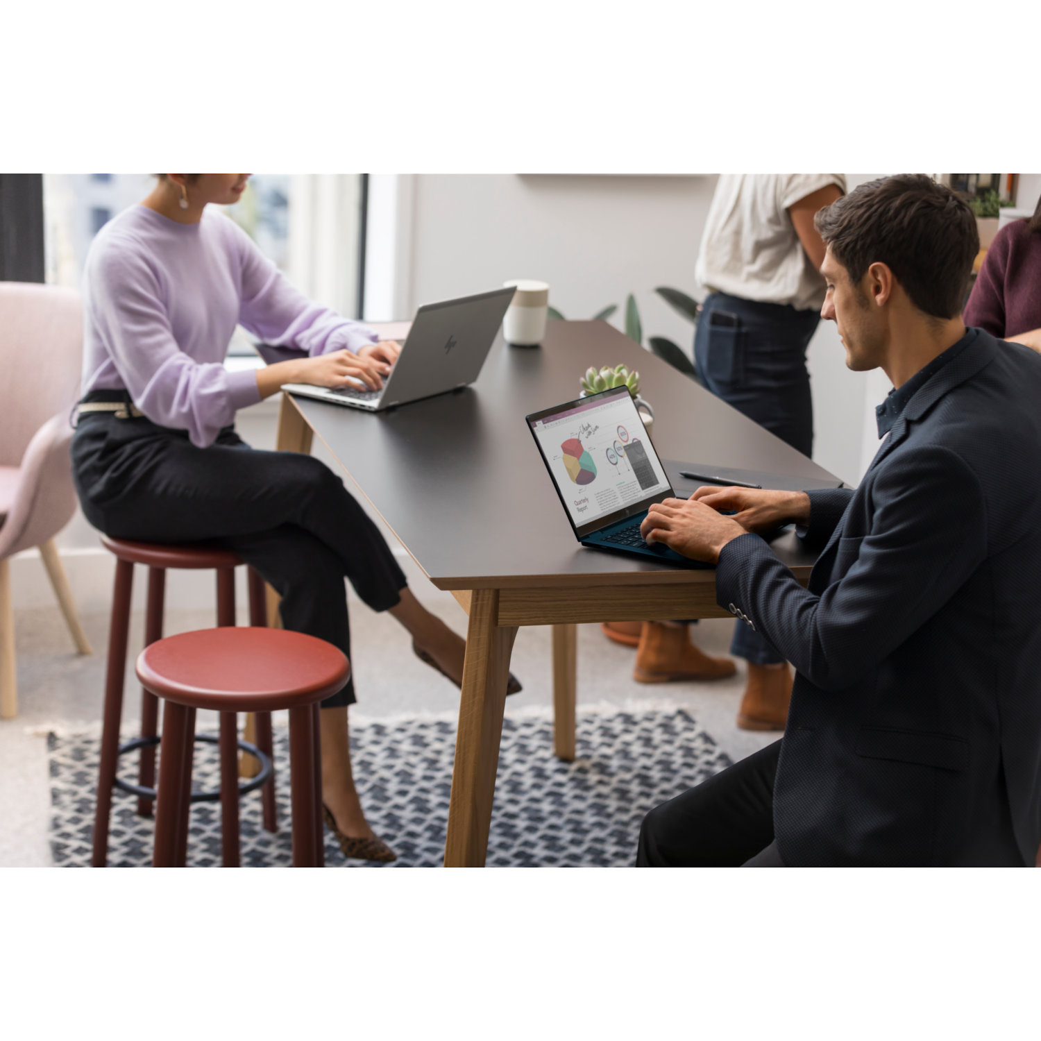 People work on laptops at a table. A woman wears a lavender sweater. A man in a suit jacket. A red stool is in the foreground.
