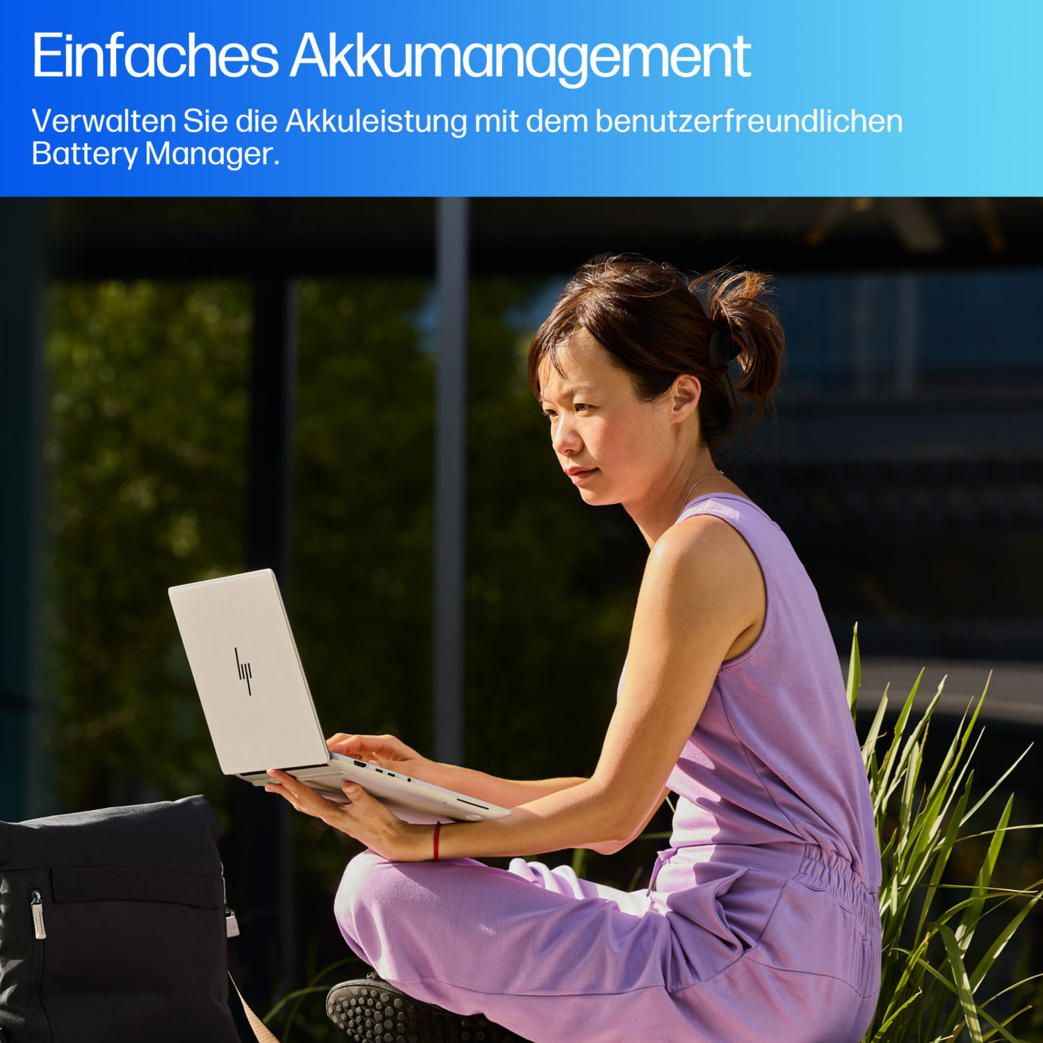 A woman in a purple outfit sits outdoors, using a laptop. A black bag is beside her. Green plants and a blurred building are in the background.